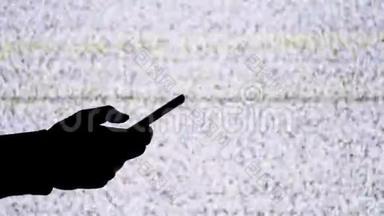 一只手拿着智能手机在电视屏幕背景上的剪影，带有白色的静态噪音